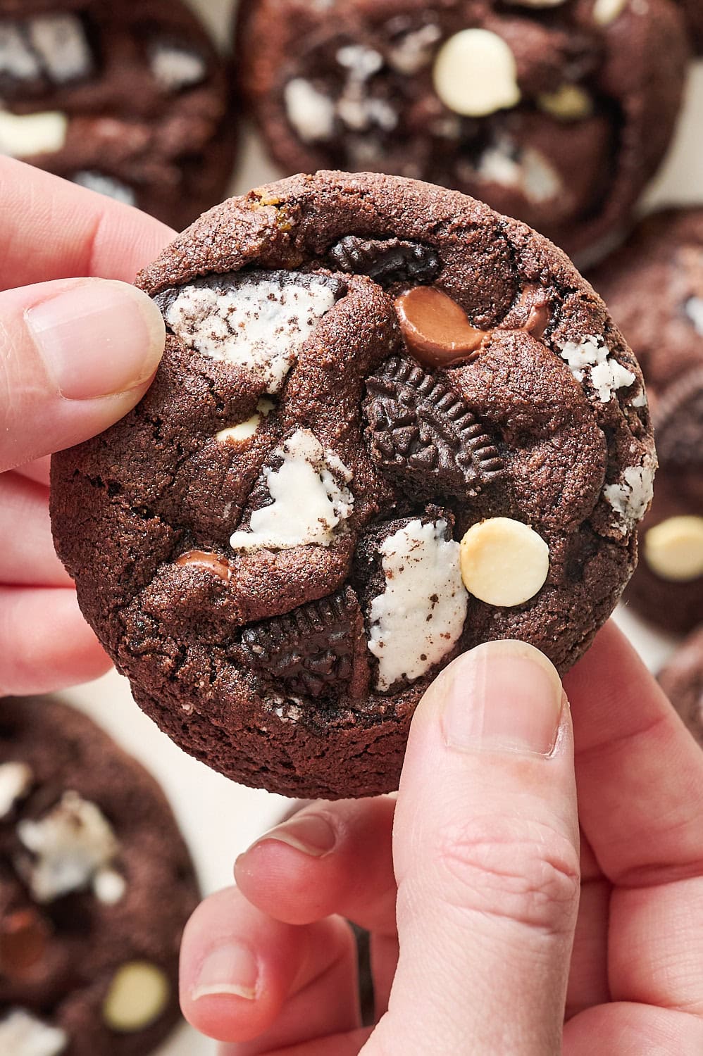 hands holding a chocolate cookies & cream cookie, with more cookies on the counter in the background