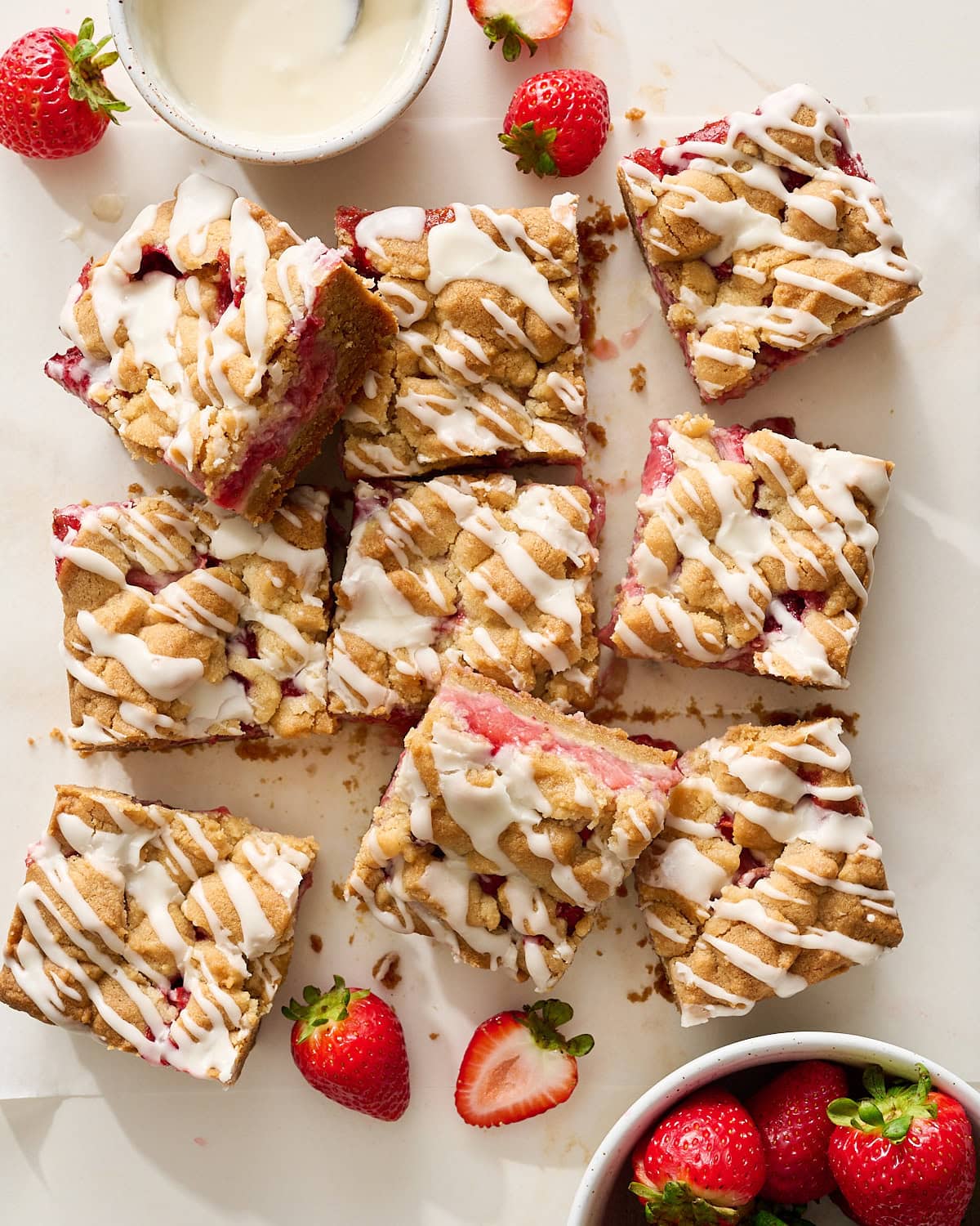 overhead of strawberry cobbler bars on a marble surface with more strawberries and the bowl of icing
