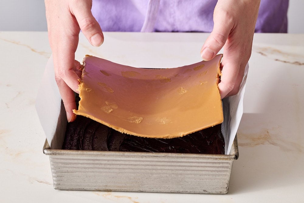 two hands placing solid sheet of Biscoff spread onto brownie batter in pan