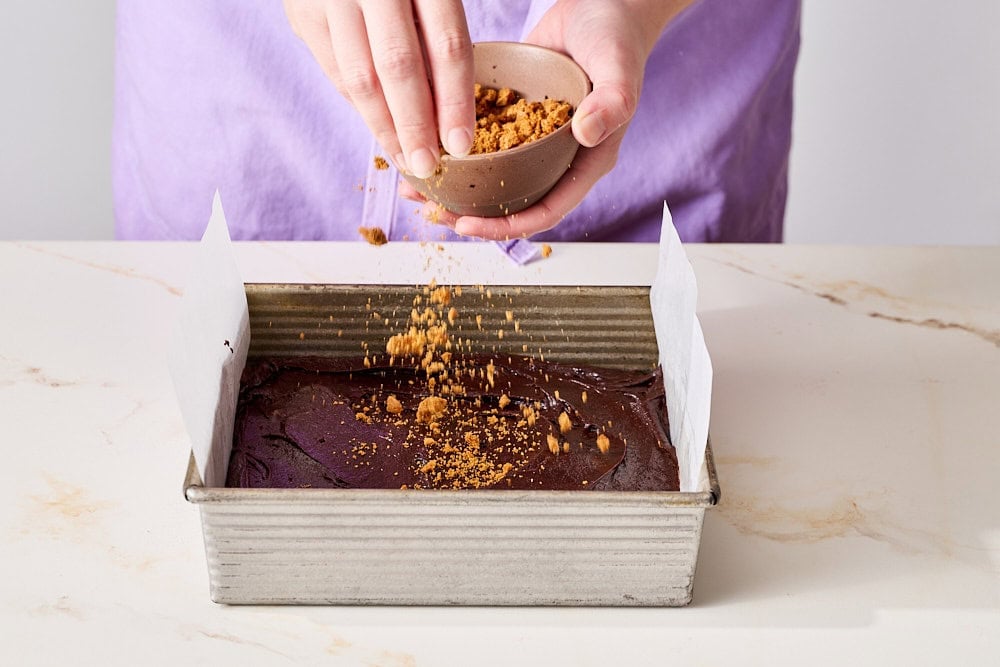 hands sprinkling brownie batter in pan with crushed Biscoff cookies