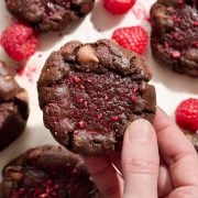 One hand holding a thick chocolate raspberry truffle cookie, with more cookies and raspberries on the background surface