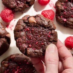 One hand holding a thick chocolate raspberry truffle cookie, with more cookies and raspberries on the background surface