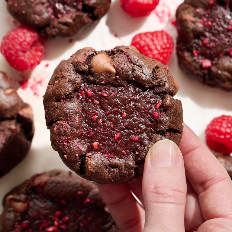One hand holding a thick chocolate raspberry truffle cookie, with more cookies and raspberries on the background surface