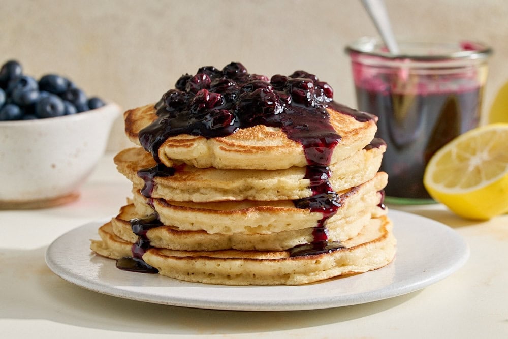 blueberry sauce on a stack of pancakes with a lemon, more blueberries, and extra sauce in the background