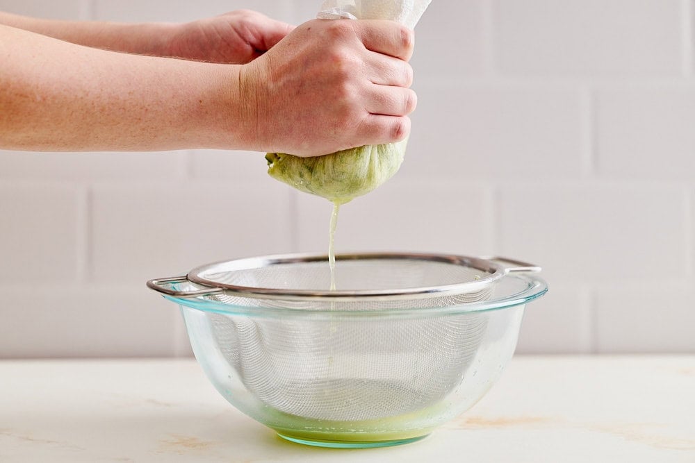 two hands squeezing the excess moisture out of the shredded zucchini and apple into the glass bowl