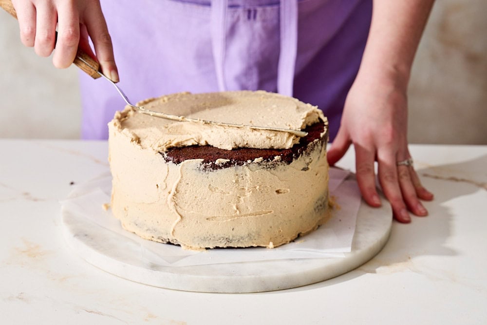 Tessa spreading peanut butter frosting on an assembled chocolate cake with an offset spatula