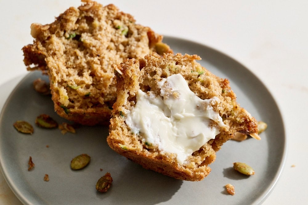a zucchini apple muffin on a small gray plate, cut in half, with butter smeared on one half