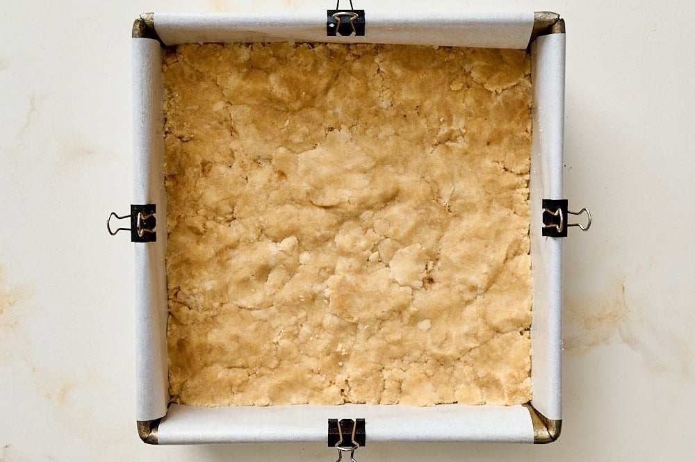 overhead view of shortbread dough pressed into bottom of 8-inch square metal baking pan lined with parchment