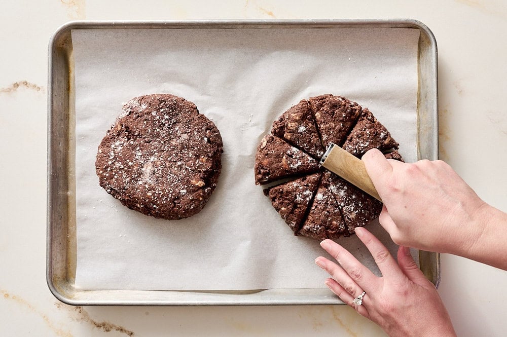 overhead view of two disks of scone dough with a hand cutting one disk into triangles with a bench scraper