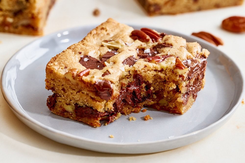 close view of a single bourbon pecan chocolate chunk blondie on a plate with a bite taken out to show chewy interior
