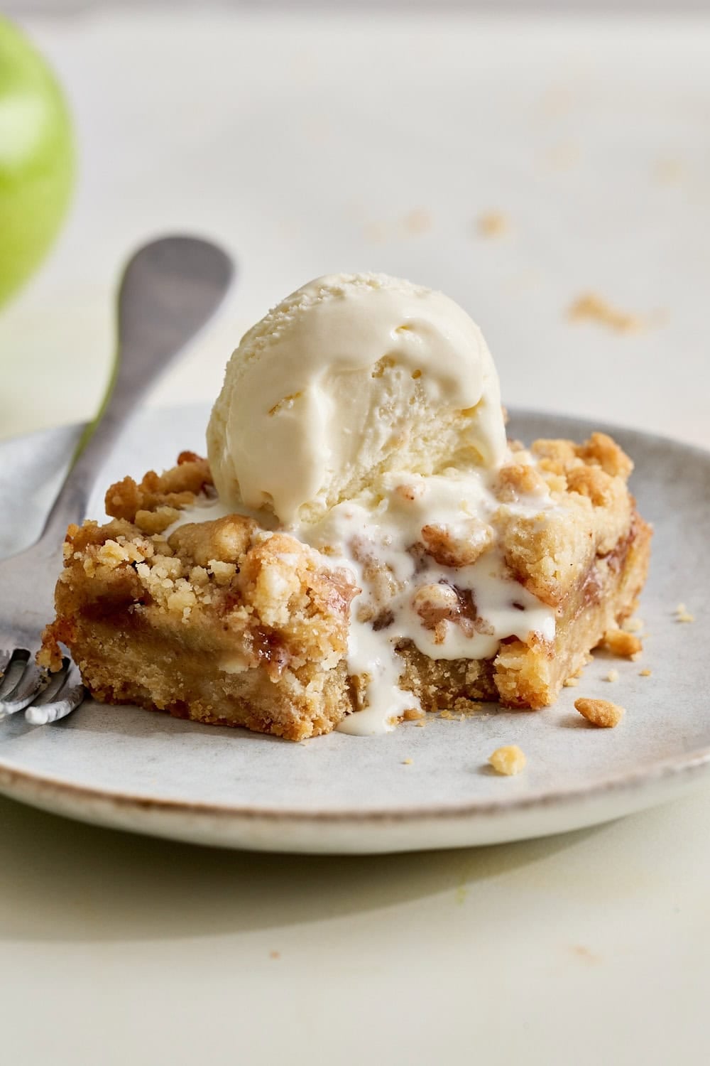 an apple pie bar on a small plate with a scoop of melt-y vanilla ice cream on top and a forkful taken out