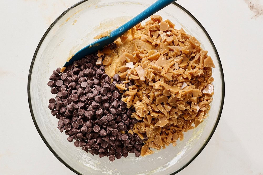 chocolate chips and toffee bits poured over peanut butter cookie dough in glass mixing bowl