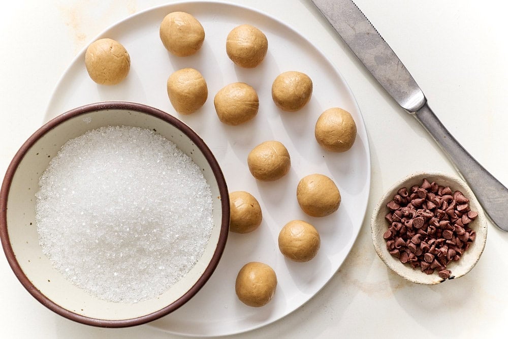 overhead view of peanut butter mixture rolled into balls with bowl of mini chocolate chips and a bowl of sparkling sugar