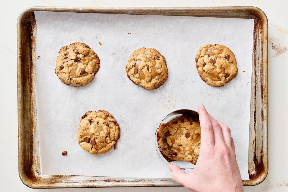 overhead view of freshly baked cookies with a hand using a round cookie cutter to re-shape into perfect circles