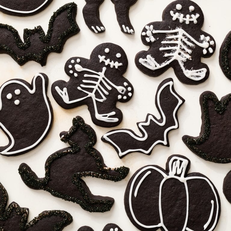 overhead view of many black cocoa Halloween sugar cookies decorated into various shapes including skeletons, ghosts, pumpkins, bats, and cats