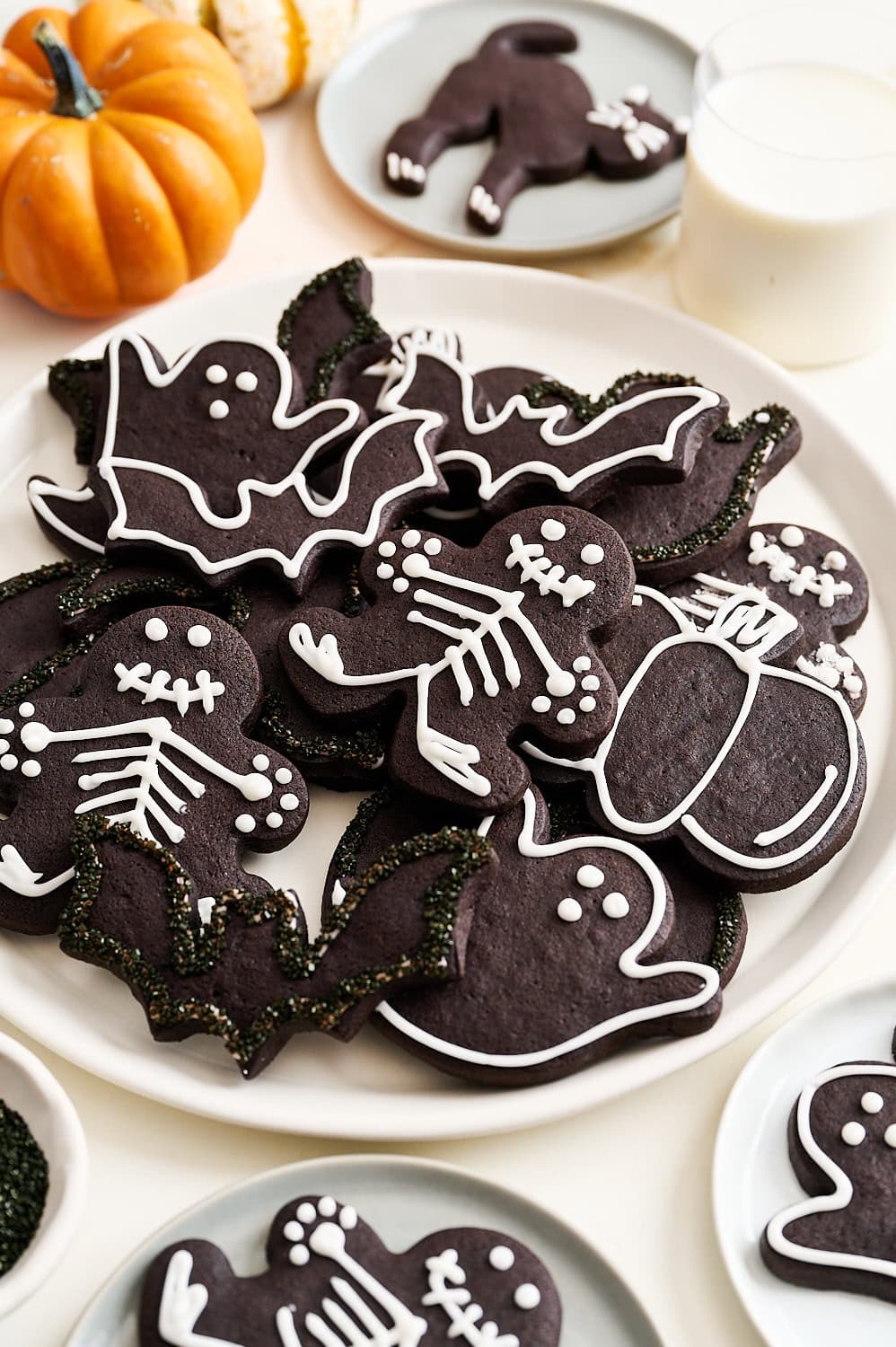 many black cocoa Halloween sugar cookies on a white plate decorated into various shapes including skeletons, ghosts, pumpkins, bats, and cats