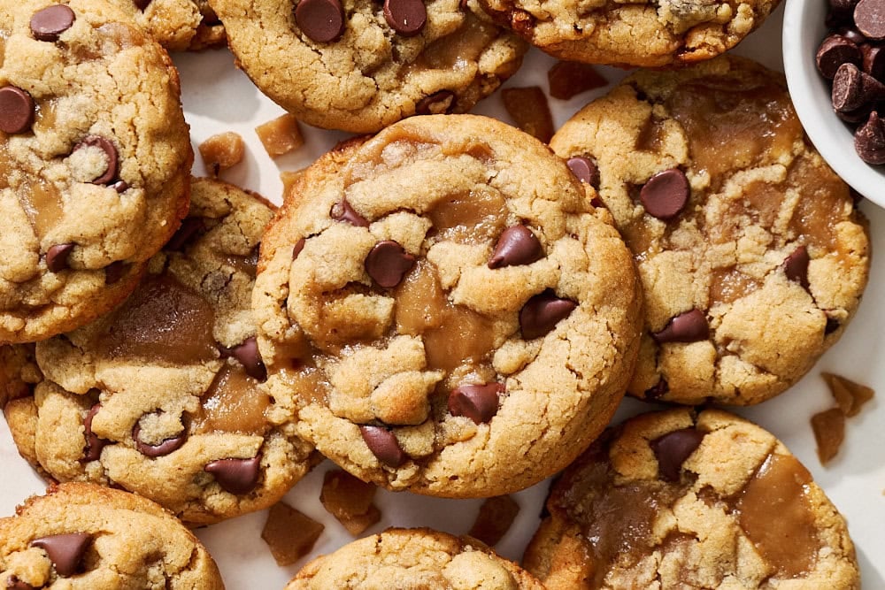 many thick and chewy peanut butter toffee cookies on a marble counter next to a bowl of extra chocolate chips